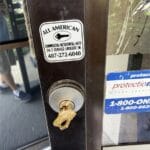 A metal door lock with a key inserted and an All American locksmith sticker visible on the glass door.