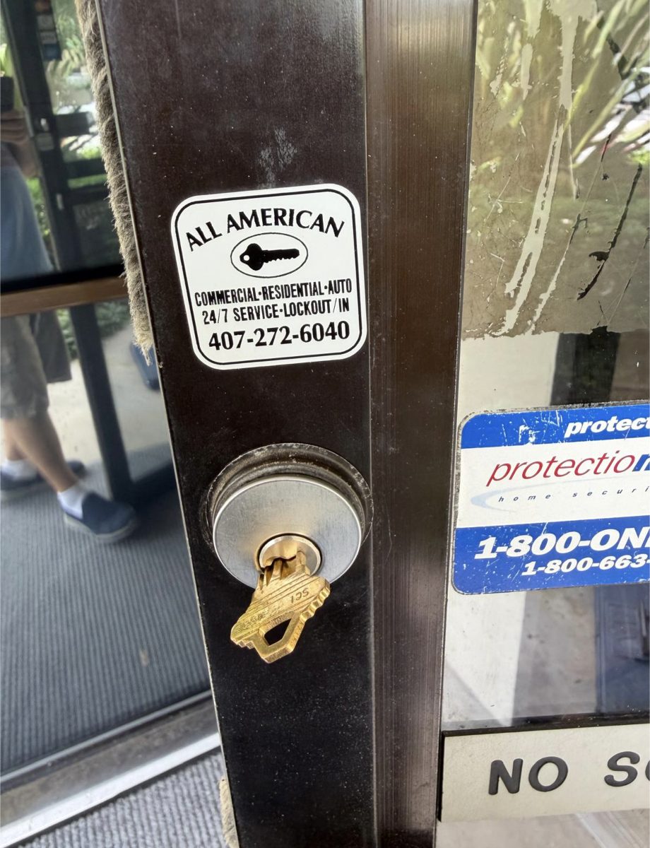 A metal door lock with a key inserted and an All American locksmith sticker visible on the glass door.